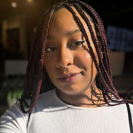 Pearl with smedium purple braids smiling sweetly at you in warm lighting with a white shirt on.